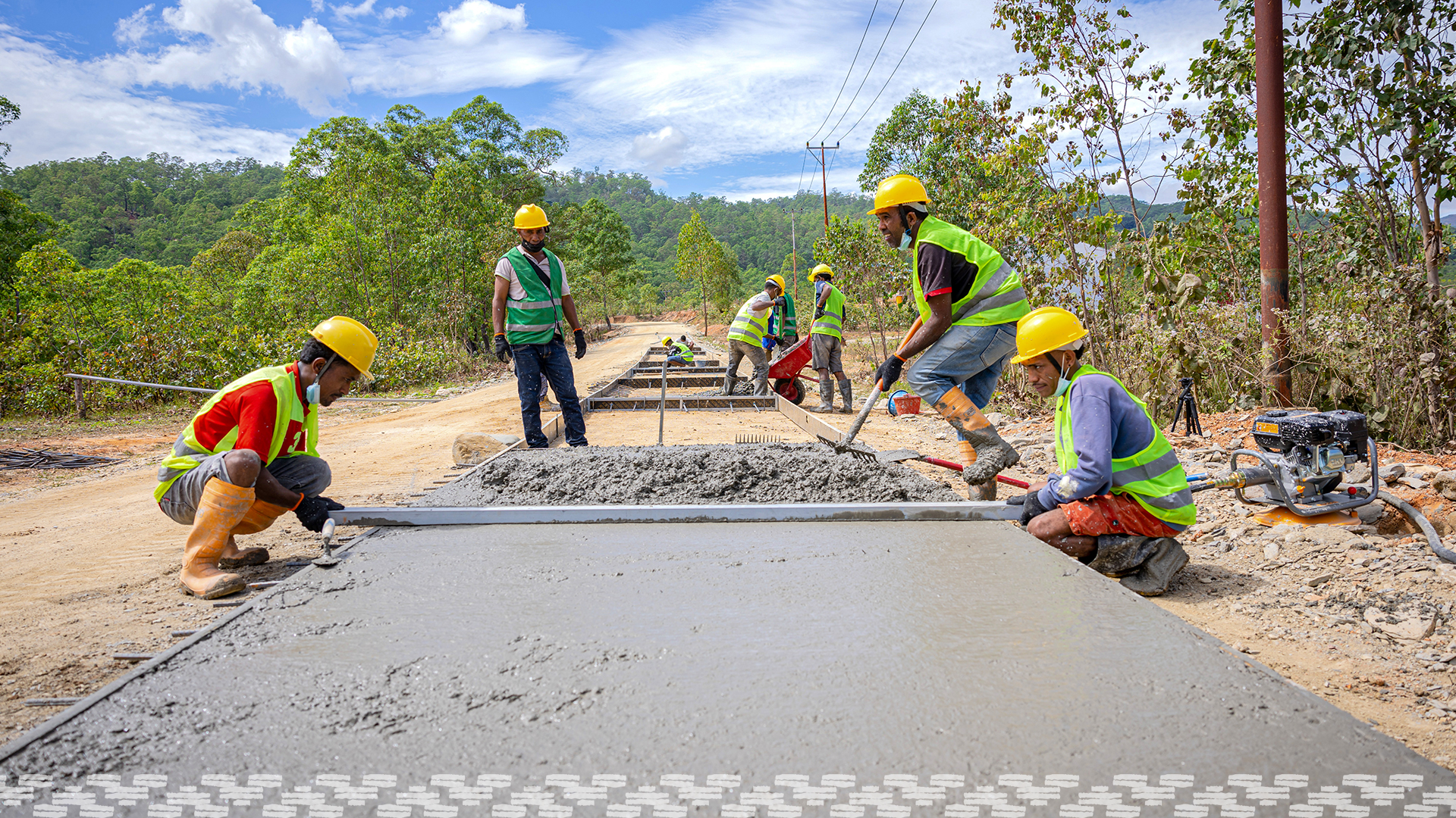 A group of road workers in high visibility clothing and hard hats are smoothing wet cement as they build a road in a rural setting in Timor-Leste.