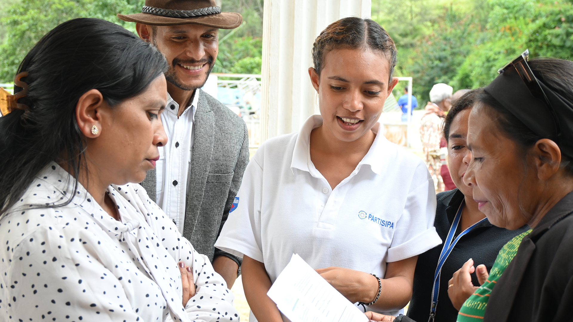 A young woman in a PARTISIPA t-shirt is standing with a group of men and women, they are smiling as they discuss something.